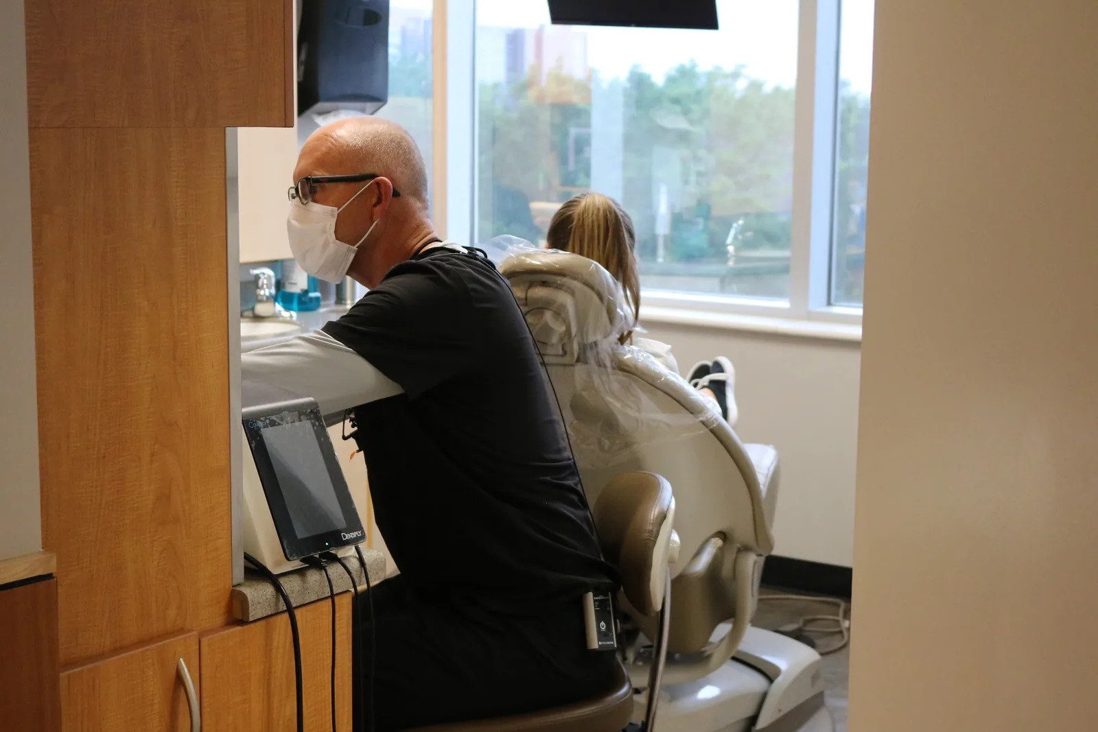 A dental professional wearing a face mask seated beside a patient in a dental chair.